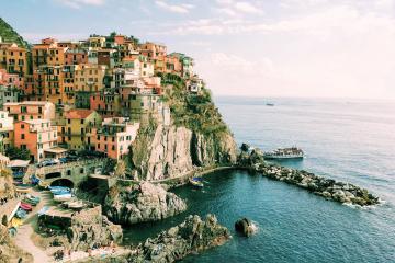 A colorful village built into a steep cliffside overlooking the sea, with a small harbor and boats visible in the water below.
