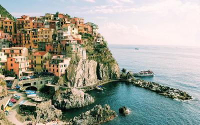 A colorful village built into a steep cliffside overlooking the sea, with a small harbor and boats visible in the water below.