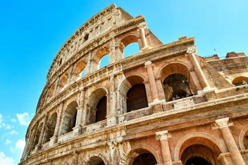 The image shows the exterior of the Colosseum in Rome, Italy, featuring its iconic arched stone architecture against a clear blue sky.
