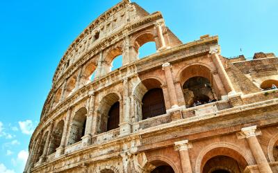 The image shows the exterior of the Colosseum in Rome, Italy, featuring its iconic arched stone architecture against a clear blue sky.