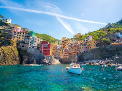 A scenic view of colorful houses built into a steep cliffside overlooking turquoise water with boats and swimmers, characteristic of the Cinque Terre villages in Italy.