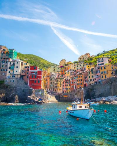 A scenic view of colorful houses built into a steep cliffside overlooking turquoise water with boats and swimmers, characteristic of the Cinque Terre villages in Italy.
