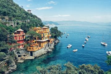 A coastal scene featuring colorful villas built into a steep, green cliffside overlooking a bay with numerous sailboats and yachts anchored in clear blue water.