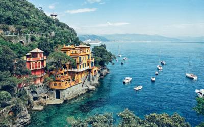 A coastal scene featuring colorful villas built into a steep, green cliffside overlooking a bay with numerous sailboats and yachts anchored in clear blue water.