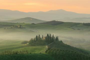 A misty Tuscan landscape at sunrise or sunset featuring rolling green hills, a cluster of cypress trees, and a small farmhouse on a hilltop, with distant mountains under a soft orange sky.