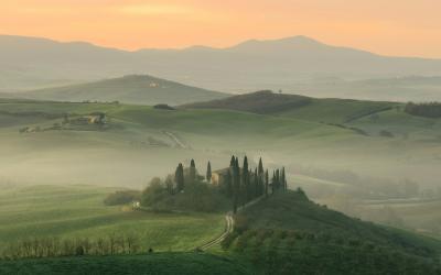 A misty Tuscan landscape at sunrise or sunset featuring rolling green hills, a cluster of cypress trees, and a small farmhouse on a hilltop, with distant mountains under a soft orange sky.