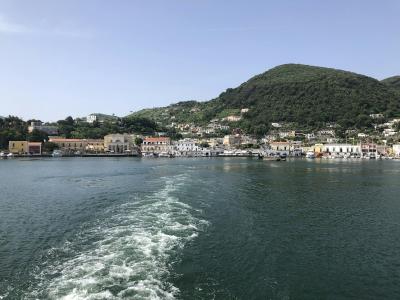 The image shows the wake of a ferry boat on the water, with a coastal town and a green hill in the background under a clear sky.