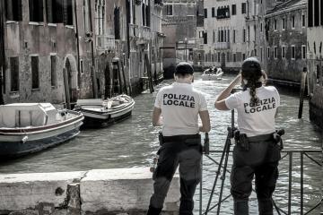 Two uniformed officers with "POLIZIA LOCALE" on their backs stand on a canal bank in Venice, Italy, looking out over the water where boats are moored.