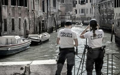 Two uniformed officers with "POLIZIA LOCALE" on their backs stand on a canal bank in Venice, Italy, looking out over the water where boats are moored.