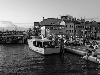 A black-and-white photograph of a harbor scene featuring a white passenger boat docked at a concrete pier, with other fishing boats moored in the background and multi-story buildings lining the waterfront under a twilight sky.