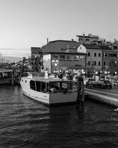 A black-and-white photograph of a harbor scene featuring a white passenger boat docked at a concrete pier, with other fishing boats moored in the background and multi-story buildings lining the waterfront under a twilight sky.