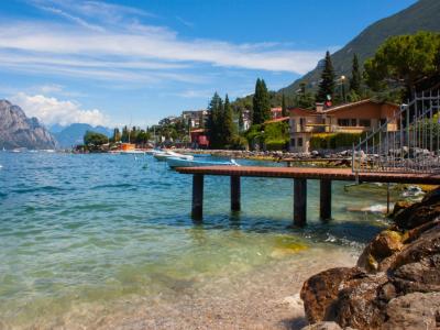 A sunny lakeside scene featuring a wooden pier extending into clear turquoise water, with a rocky shoreline in the foreground, boats moored nearby, and a hillside town with terracotta-roofed buildings and greenery under a blue sky.