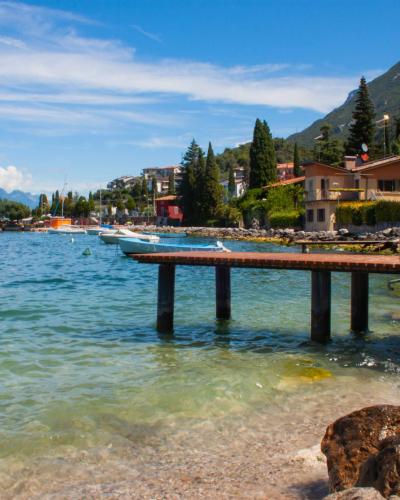 A sunny lakeside scene featuring a wooden pier extending into clear turquoise water, with a rocky shoreline in the foreground, boats moored nearby, and a hillside town with terracotta-roofed buildings and greenery under a blue sky.