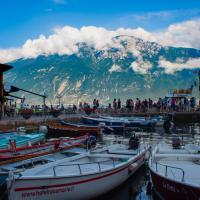 A harbor scene with several small boats moored in the foreground, a pier with people standing on it, and a large mountain range with snow-capped peaks and clouds in the background under a blue sky.
