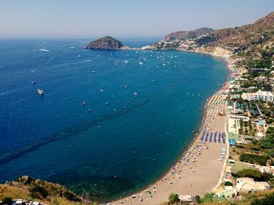 A high-angle panoramic view of a long, curved sandy beach lined with colorful umbrellas and sunbathers, backed by multi-story hotels and buildings, with a prominent rocky islet offshore and hills rising behind the town under a clear blue sky.