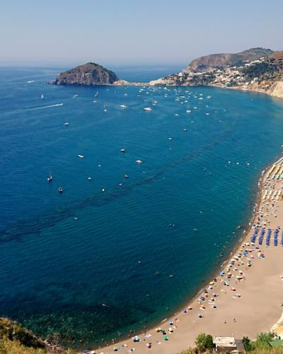 A high-angle panoramic view of a long, curved sandy beach lined with colorful umbrellas and sunbathers, backed by multi-story hotels and buildings, with a prominent rocky islet offshore and hills rising behind the town under a clear blue sky.