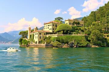 A large, historic villa complex with terraced gardens situated on a rocky promontory extending into a lake, with a white motorboat passing in the foreground and mountains visible in the distance.