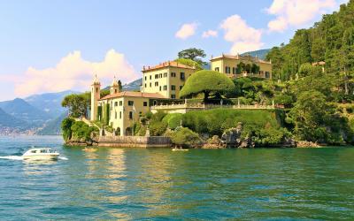 A large, historic villa complex with terraced gardens situated on a rocky promontory extending into a lake, with a white motorboat passing in the foreground and mountains visible in the distance.