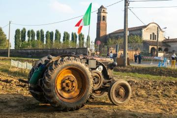 A vintage green tractor with large rear wheels sits in a muddy field in the foreground, with an Italian flag on a pole, a stone church with a bell tower, and people gathered near a building in the background under a clear sky.