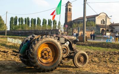 A vintage green tractor with large rear wheels sits in a muddy field in the foreground, with an Italian flag on a pole, a stone church with a bell tower, and people gathered near a building in the background under a clear sky.