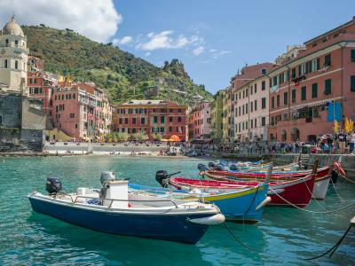 A sunlit coastal harbor scene featuring a small white boat moored in turquoise water, with colorful multi-story buildings densely packed along a steep cliffside under a blue sky with contrails.