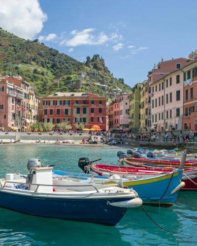 A sunlit coastal harbor scene featuring a small white boat moored in turquoise water, with colorful multi-story buildings densely packed along a steep cliffside under a blue sky with contrails.