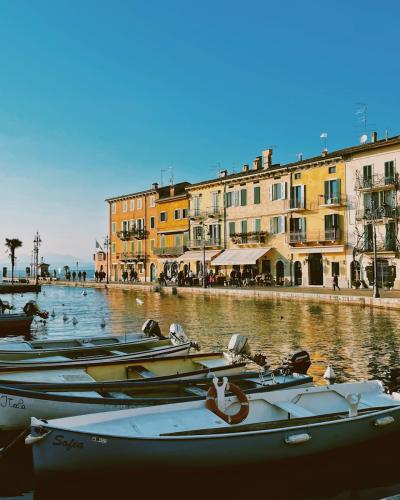 The image shows a row of small white motorboats moored in calm water along a stone quay, with a backdrop of colorful multi-story buildings and a clear blue sky.