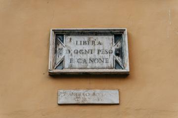 A weathered stone plaque mounted on a yellow stucco wall, inscribed with the Italian text "LIBERA DA OGNI PESO E CANONE" (Free from every weight and fee/tax), with a smaller, illegible plaque below it.
