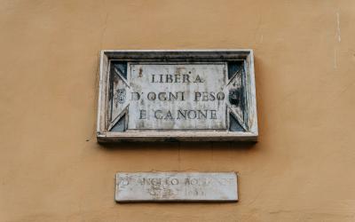 A weathered stone plaque mounted on a yellow stucco wall, inscribed with the Italian text "LIBERA DA OGNI PESO E CANONE" (Free from every weight and fee/tax), with a smaller, illegible plaque below it.