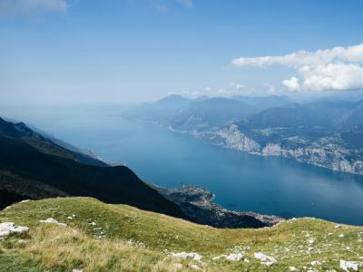 A high-angle landscape view of a large blue lake surrounded by steep mountains, with a grassy foreground and a town visible on the shoreline.
