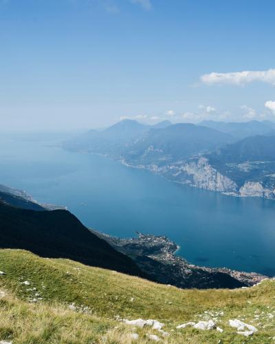 A high-angle landscape view of a large blue lake surrounded by steep mountains, with a grassy foreground and a town visible on the shoreline.