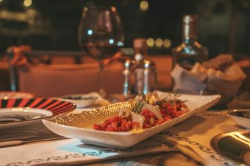 A close-up of a rectangular white serving dish with gold trim containing a colorful appetizer or antipasto, set on a table with wine glasses, condiments, and other dining ware in a dimly lit restaurant.