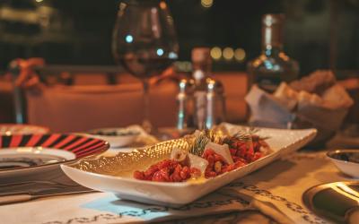 A close-up of a rectangular white serving dish with gold trim containing a colorful appetizer or antipasto, set on a table with wine glasses, condiments, and other dining ware in a dimly lit restaurant.