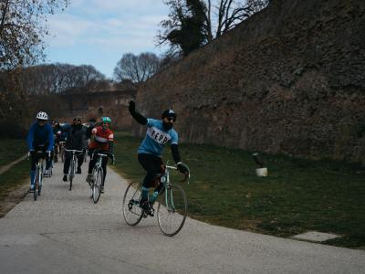 A group of cyclists rides on a paved path alongside a tall, ancient brick wall under an overcast sky, with one cyclist in the foreground raising an arm.