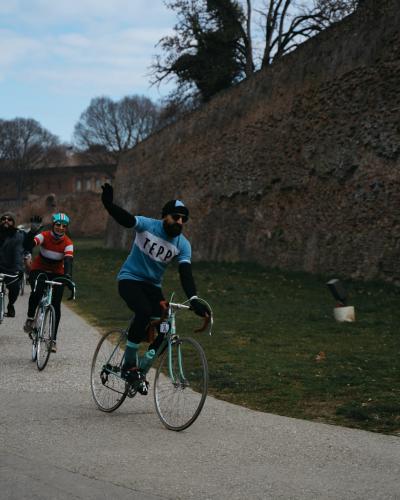 A group of cyclists rides on a paved path alongside a tall, ancient brick wall under an overcast sky, with one cyclist in the foreground raising an arm.