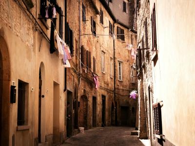 A narrow, sunlit cobblestone street in an old European town, featuring tall, weathered buildings with arched doorways, shuttered windows, and laundry hanging from balconies.