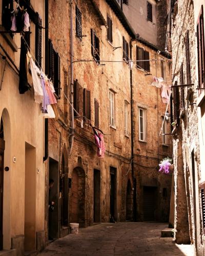 A narrow, sunlit cobblestone street in an old European town, featuring tall, weathered buildings with arched doorways, shuttered windows, and laundry hanging from balconies.