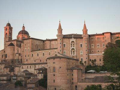 A large, historic Renaissance palace complex with multiple towers, a central dome, and terracotta-colored stone walls, viewed from a lower vantage point with rooftops and trees in the foreground under a soft sky.