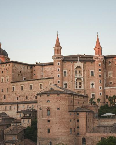 A large, historic Renaissance palace complex with multiple towers, a central dome, and terracotta-colored stone walls, viewed from a lower vantage point with rooftops and trees in the foreground under a soft sky.