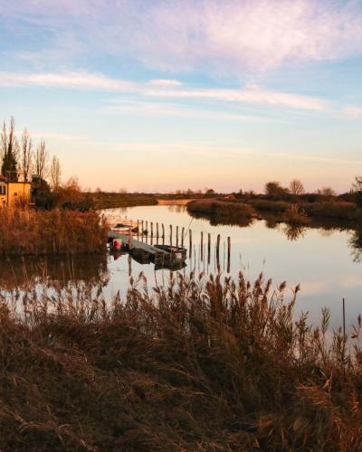 A small boat is moored in a calm, narrow waterway surrounded by tall reeds and grasses, with a yellow building visible on the left bank under a twilight sky.