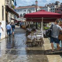 A flooded outdoor cafe area in Venice with tables and chairs partially submerged in water, red umbrellas, pedestrians walking on elevated walkways, and buildings lining a canal or square.