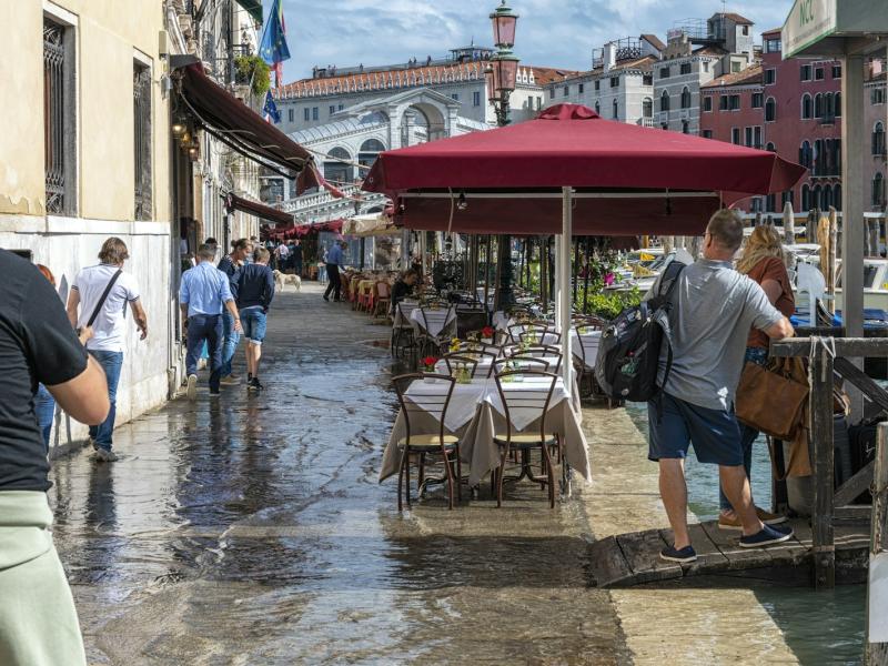 A flooded outdoor cafe area in Venice with tables and chairs partially submerged in water, red umbrellas, pedestrians walking on elevated walkways, and buildings lining a canal or square.