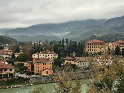 A wide-angle view of a river flowing through a town with terracotta-roofed buildings, a stone embankment, and misty hills in the background; no visible signage or specific landmarks like the Alpini Bridge are clearly identifiable.
