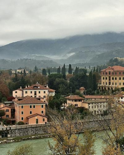 A wide-angle view of a river flowing through a town with terracotta-roofed buildings, a stone embankment, and misty hills in the background; no visible signage or specific landmarks like the Alpini Bridge are clearly identifiable.