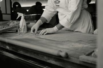 A person in a chef's coat and gloves is working with dough on a wooden counter, with a spray bottle and rolling pin visible.