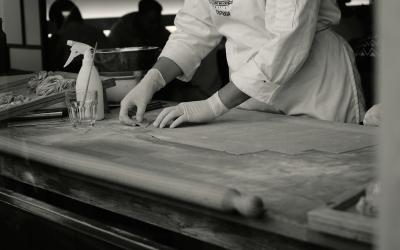 A person in a chef's coat and gloves is working with dough on a wooden counter, with a spray bottle and rolling pin visible.