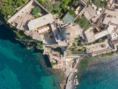 An aerial view of a coastal town with terracotta-roofed buildings, a prominent domed church, and a harbor with boats, situated on a peninsula surrounded by turquoise water.