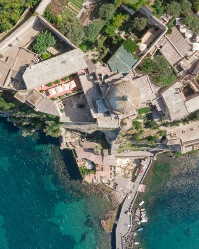An aerial view of a coastal town with terracotta-roofed buildings, a prominent domed church, and a harbor with boats, situated on a peninsula surrounded by turquoise water.