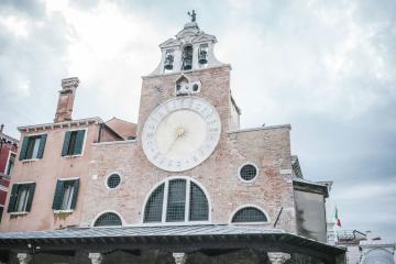 A low-angle shot of a pinkish-brick clock tower with a large white clock face, a bell gable, and an arched arcade below, set against a cloudy sky.