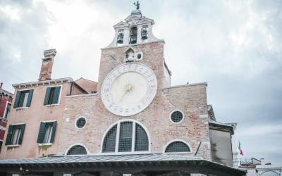 A low-angle shot of a pinkish-brick clock tower with a large white clock face, a bell gable, and an arched arcade below, set against a cloudy sky.